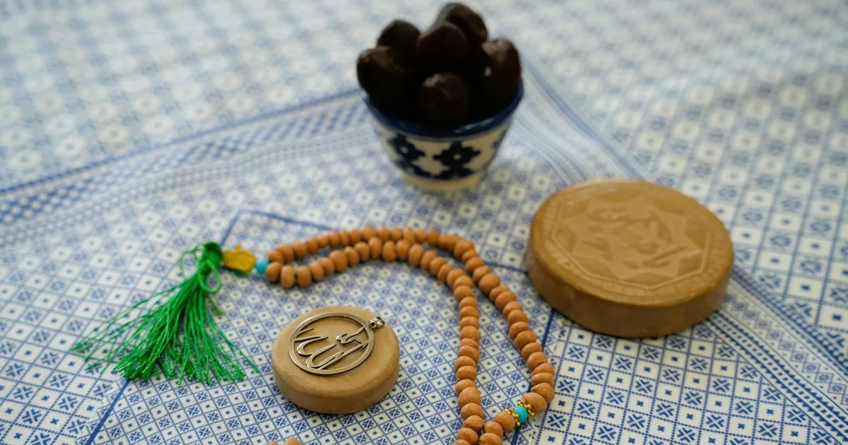 Wooden prayer beads with a green tassel and an Allah pendant beside a small bowl of dates on a blue patterned prayer mat