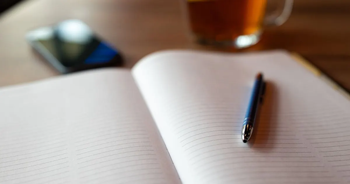 An open lined notebook with a blue pen, a glass of tea and a phone on a wooden table, ready for planning together