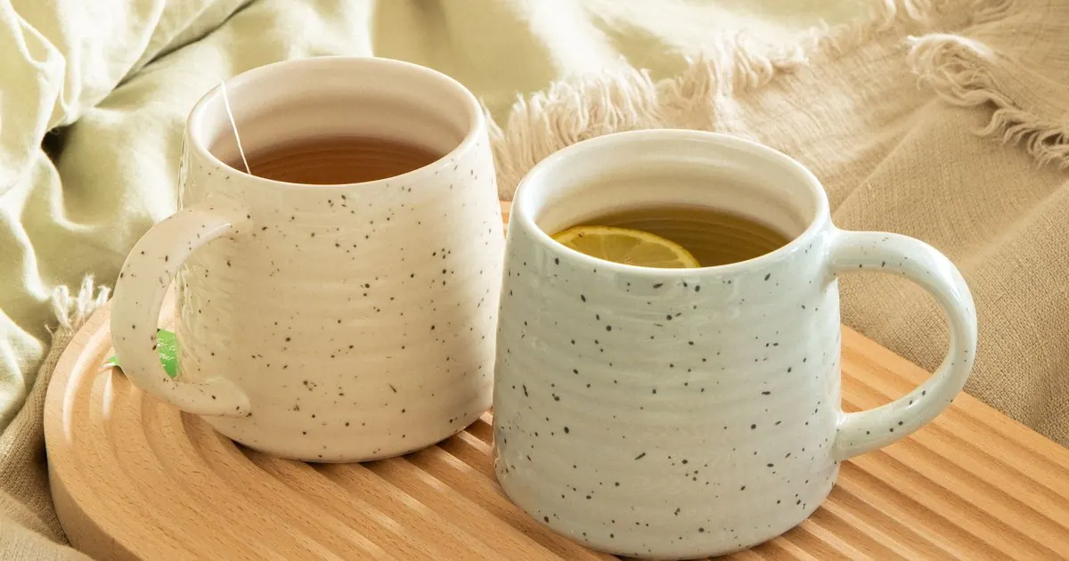 Two speckled ceramic mugs of tea side by side on a wooden tray over a soft linen blanket