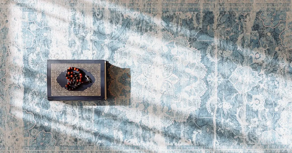 A closed Quran with prayer beads on a patterned prayer rug in soft sunlight